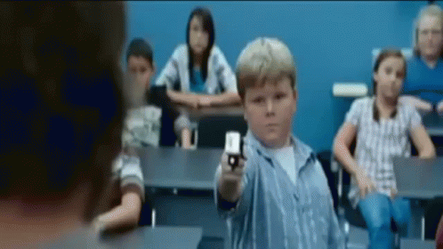 A Young Boy Is Pointing A Gun In A Classroom With Other Children GIF