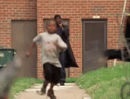 A Young Boy Is Running Down A Sidewalk In Front Of A Brick Building While A Man Watches . GIF