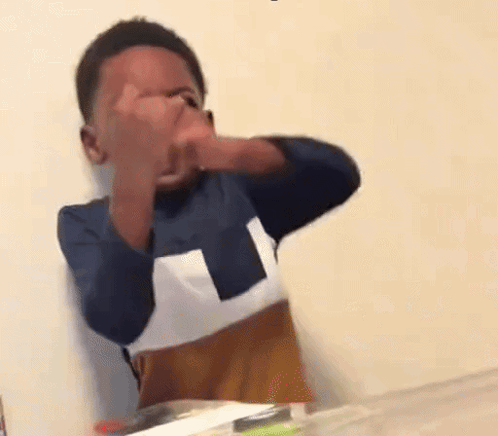 A Young Boy Is Sitting At A Table Covering His Face With His Hands . GIF