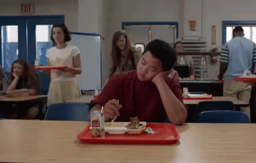 A Young Boy Is Sitting At A Table In A School Cafeteria Eating A Meal . GIF