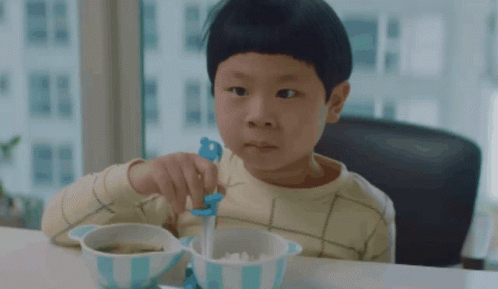 A Young Boy Is Sitting At A Table With A Bowl Of Food And A Blue Spoon GIF