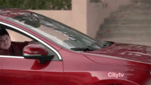 A Young Boy Is Sitting In A Red Car With A Citytv Sticker On The Side . GIF