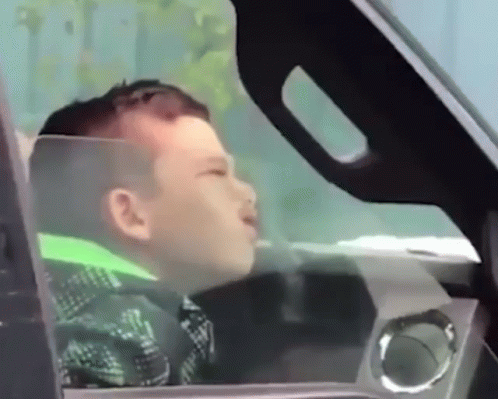 A Young Boy Is Sitting In The Driver 'S Seat Of A Car Looking Out The Window GIF