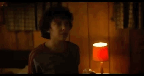 A Young Boy Is Standing In A Room With A Lamp And A Wooden Wall . GIF
