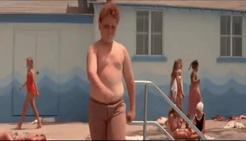 A Young Boy Is Standing In Front Of A Swimming Pool While A Group Of Children Are Playing In The Water . GIF