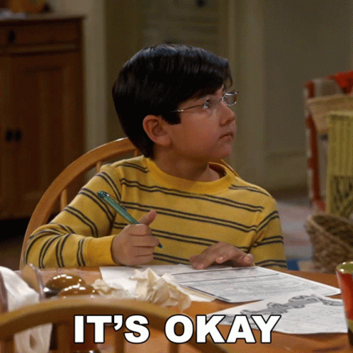 A Young Boy Sits At A Table With A Pen In His Hand And The Words " It 'S Okay " Written Below Him GIF