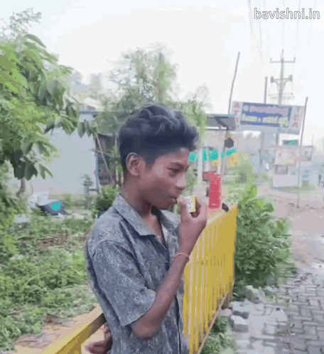 A Young Boy Smoking A Cigarette In Front Of A Yellow Fence With The Website Bavishni.in Written On The Bottom GIF