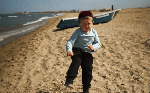 A Young Boy Stands On A Beach With A Boat In The Background GIF