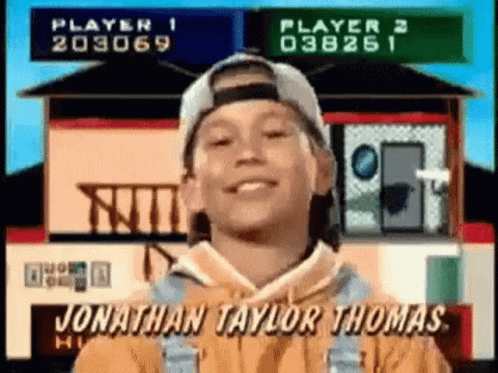 A Young Boy Wearing A Baseball Cap And Overalls Is Smiling In Front Of A House . GIF