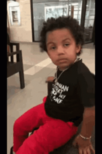 A Young Boy Wearing A Black Shirt And Red Pants Is Sitting On A Chair And Looking At The Camera . GIF