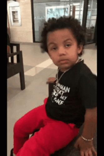 A Young Boy Wearing A Black Shirt That Says ' Woman ' On It Is Sitting On A Chair GIF