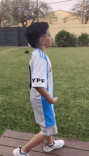 A Young Boy Wearing A Blue And White Ypf Jersey Walks Across A Wooden Deck GIF