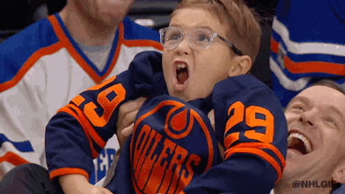A Young Boy Wearing A Blue Oilers Jersey Is Being Held By A Man GIF