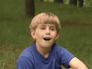 A Young Boy Wearing A Blue Shirt Is Sitting In The Grass With His Mouth Open GIF