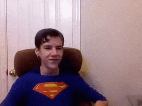 A Young Boy Wearing A Blue Superman Shirt Is Sitting In A Chair . GIF