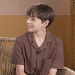 A Young Boy Wearing A Brown Shirt Is Sitting On A Chair . GIF