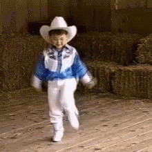 A Young Boy Wearing A Cowboy Hat And A Blue Jacket Is Walking On A Wooden Floor . GIF