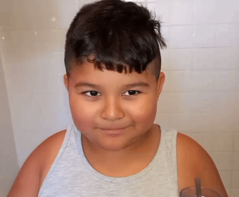 A Young Boy Wearing A Grey Tank Top Is Smiling And Holding A Glass GIF