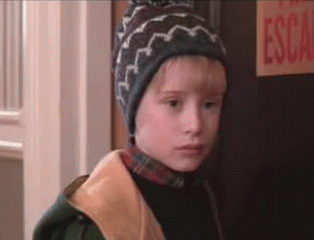A Young Boy Wearing A Hat And A Jacket Is Standing In Front Of A Door . GIF