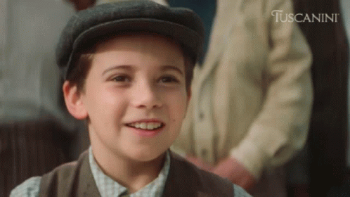 A Young Boy Wearing A Hat Is Smiling In Front Of A Sign That Says Tuscanini GIF
