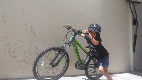 A Young Boy Wearing A Helmet Is Standing Next To A Green Bicycle GIF