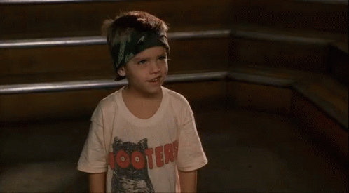 A Young Boy Wearing A Hooters T-Shirt And A Headband Is Standing In Front Of Some Stairs . GIF
