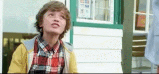 A Young Boy Wearing A Plaid Shirt And A Yellow Jacket Is Standing In Front Of A White Building . GIF