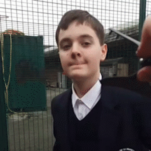A Young Boy Wearing A School Uniform Stands In Front Of A Fence GIF