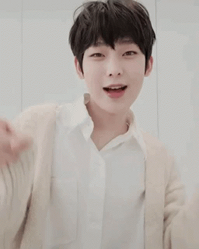 A Young Boy Wearing A White Shirt And A Beige Cardigan Is Smiling And Pointing . GIF