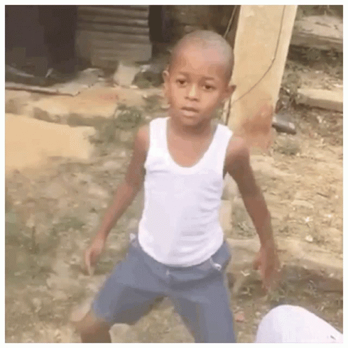 A Young Boy Wearing A White Tank Top And Blue Shorts Is Standing In A Dirt Field . GIF