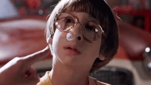 A Young Boy Wearing Glasses Is Blowing A Bubble In His Nose In Front Of A Red Car . GIF