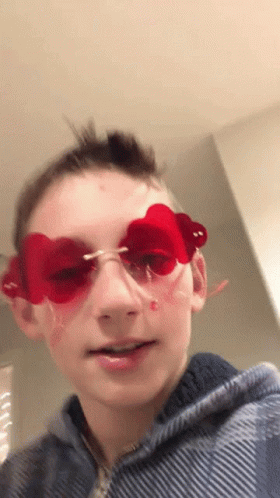 A Young Boy Wearing Red Heart Shaped Sunglasses Looks At The Camera . GIF