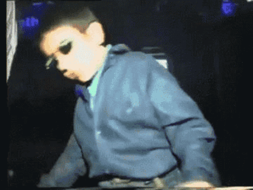A Young Boy Wearing Sunglasses And A Blue Jacket Stands In A Dark Room GIF