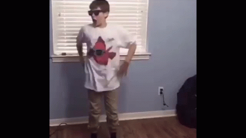 A Young Boy Wearing Sunglasses And A White Shirt Is Standing In Front Of A Window In A Room . GIF