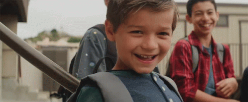 A Young Boy With A Backpack Is Smiling For The Camera While Sitting On The Stairs . GIF