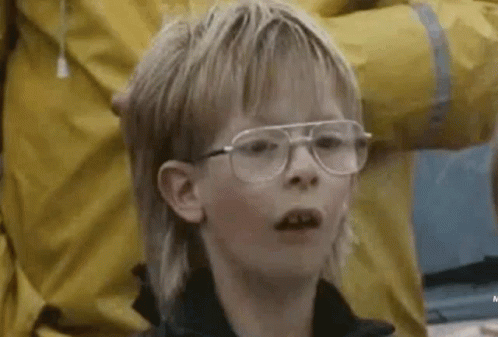 A Young Boy With A Mullet And Glasses Is Looking At The Camera . GIF