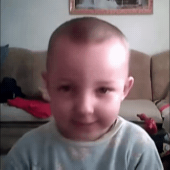 A Young Boy With A Shaved Head Is Smiling In Front Of A Couch GIF