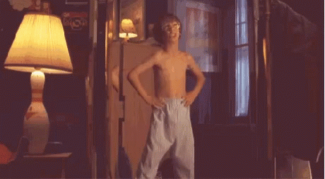 A Young Boy Without A Shirt Is Standing In A Dark Room With A Lamp . GIF