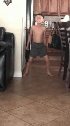 A Young Boy Without A Shirt Is Standing In A Living Room . GIF
