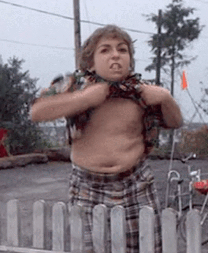 A Young Boy Without A Shirt Is Standing In Front Of A Fence And Making A Funny Face . GIF