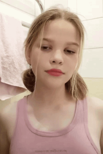A Young Girl In A Pink Tank Top Looks At The Camera With A Towel In The Background GIF