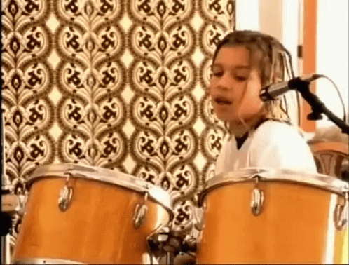 a young girl is playing drums and singing into a microphone in front of a patterned wall .