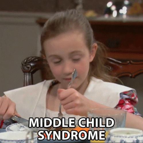 A Young Girl Is Sitting At A Table With A Fork In Her Mouth And The Words Middle Child Syndrome Written Above Her GIF