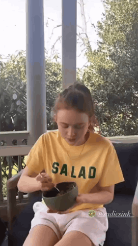 A Young Girl Is Sitting On A Porch Eating A Salad From A Bowl . GIF