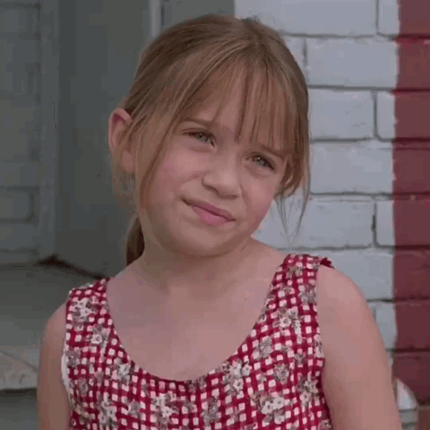 A Young Girl Wearing A Red And White Plaid Dress Stands In Front Of A Brick Wall GIF