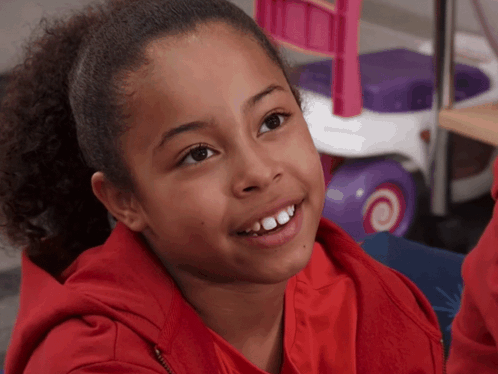 A Young Girl Wearing A Red Sweatshirt Smiles For The Camera GIF