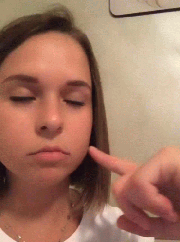 A Young Girl With Her Eyes Closed Is Pointing At Her Face GIF