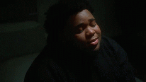 A Young Man In A Black Hoodie Is Sitting In A Dark Room With His Eyes Closed . GIF
