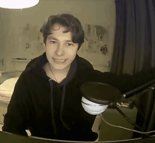 A Young Man In A Black Hoodie Stands In Front Of A Microphone And Smiles GIF