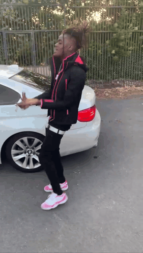 A Young Man In A Black Jacket And Pink Shoes Is Standing In Front Of A White Car GIF